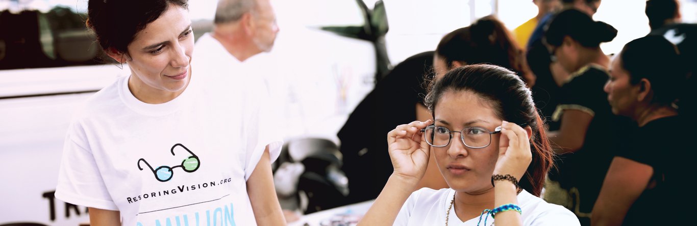 Young woman watching another woman try on new glasses at a vision event.